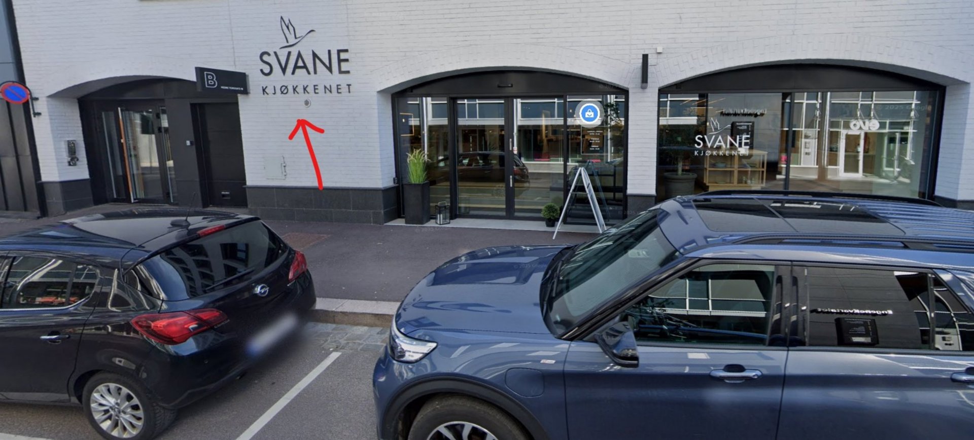 Fotografi av fasaden på en bygning med forretningslokaler (Svane Kjøkkenet), parkerte biler og en rød pil som peker på en detalj på veggen.