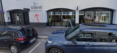 Fotografi av fasaden på en bygning med forretningslokaler (Svane Kjøkkenet), parkerte biler og en rød pil som peker på en detalj på veggen.