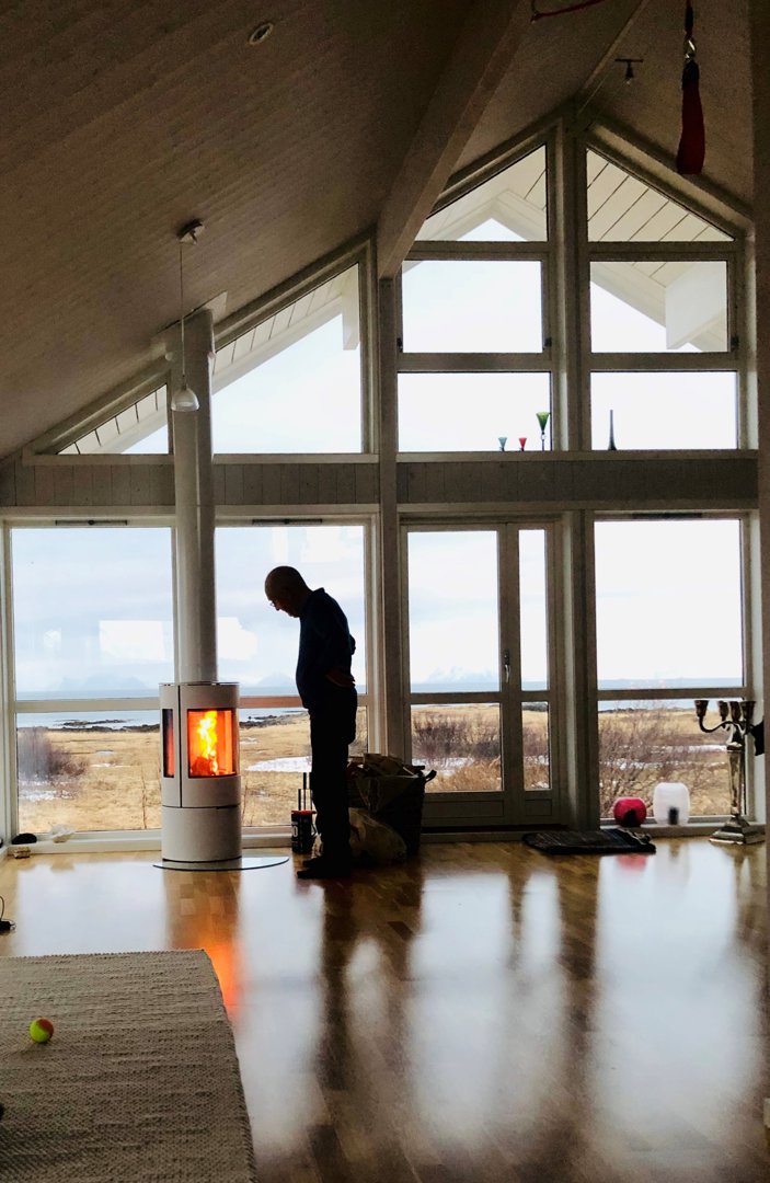 Fotografi av et stort, lys rom med store vinduer, en person ved en peis og utsikt over en kystlinje.