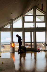 Fotografi av et stort, lys rom med store vinduer, en person ved en peis og utsikt over en kystlinje.