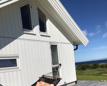 Fotografi av en hvit, paneltett bygning (sannsynligvis en hytte eller villa) med utsikt mot havet. Bildet viser fasaden, vinduer, tak og en del av en terrasse med en liggestol.