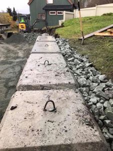 Fotografi av en byggeplass med en nybetongert vei eller gangvei i forgrunnen, en grønn husfasade i bakgrunnen og en graver i arbeid.