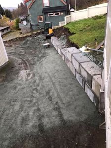 Fotografi av en byggeplass med en ny grunnmur i gang, en graver, og en eksisterende grønn husfasade i bakgrunnen.