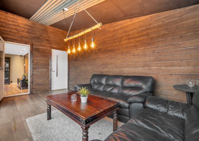 Fotografi av et innendørs rom med trepanelvegger, et svart sofa, et bord og en hengelampe.