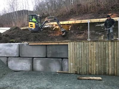 Fotografi av en byggeplass med en graver, en person og en delvis ferdigstilt fundamentmur.