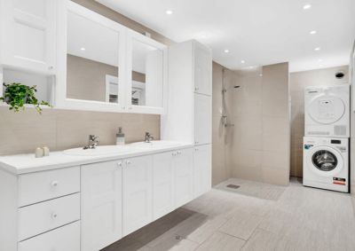 Fotografi av et moderne bad med hvite skap, to vasker, dusj og vaskemaskin.