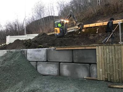 Fotografi av en byggeplass med en grøftmaskin, en person som jobber, og en betongmur under oppføring.