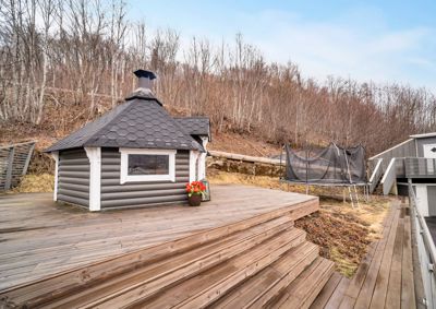 Fotografi av en liten hytte eller uthus på en treterrasse, med trampoline og skog i bakgrunnen.