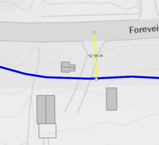 Kartutsnitt som viser tomt, veier (blå linje) og andre bygninger i nærheten. En gul målestrek indikerer en avstand på 12,99 meter mellom to punkter.