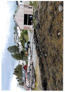 Fotografi av en tomt med en gammel, forfallelig uthus i forgrunnen, en steinmur og nabolagshus i bakgrunnen.