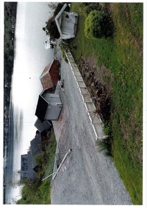 Fotografi av en rekke hytter eller båthytter langs et vann med en grusvei og en hekk.