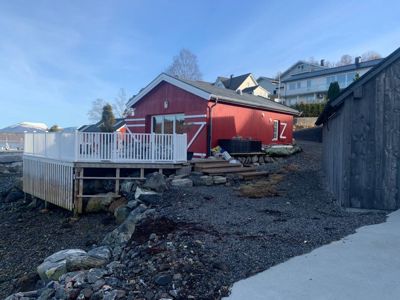 Fotografi av en rød hytte med hvit veranda og en grå uthusbygning på en steinete kysttomt.
