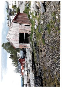 Fotografi av en gammel, rød fargebygd ved kysten med stein og grus i forgrunnen.