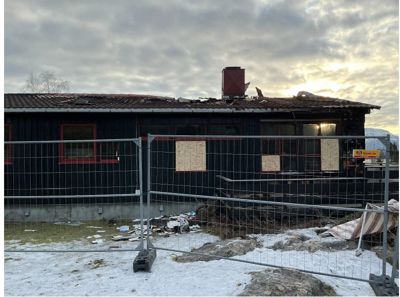 Fotografi av en mørk bygning med rødt tak, sett fra siden. Bygningen er omgitt av en metallgjerde, og det er snø på bakken. Noen vinduer er tildekket med papir.