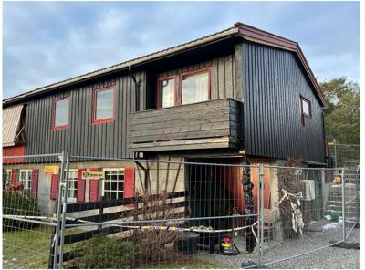 Fotografi av en eksisterende bygning med mørke trepaneler og røde vindusrammer, sett fra bakkenivå med en hekk i forgrunnen.