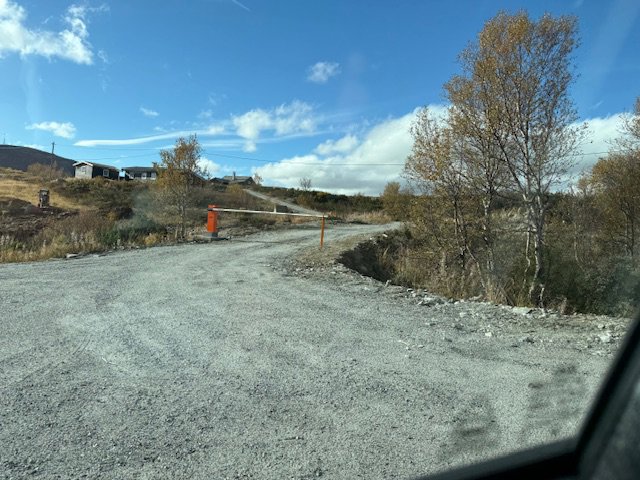 Fotografi av en grusvei som fører opp mot en bygning på en bakke, med en port i forgrunnen.