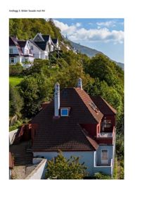 Fotografi av en bygning (hus) med rød takflate og hvite skorsteiner, sett fra en høyde. I bakgrunnen er det flere andre hus på en skråning og en fjelltopp.