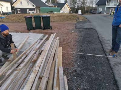 Fotografi av en byggeplass med to personer, en stakke med trevirke og en målebånd.
