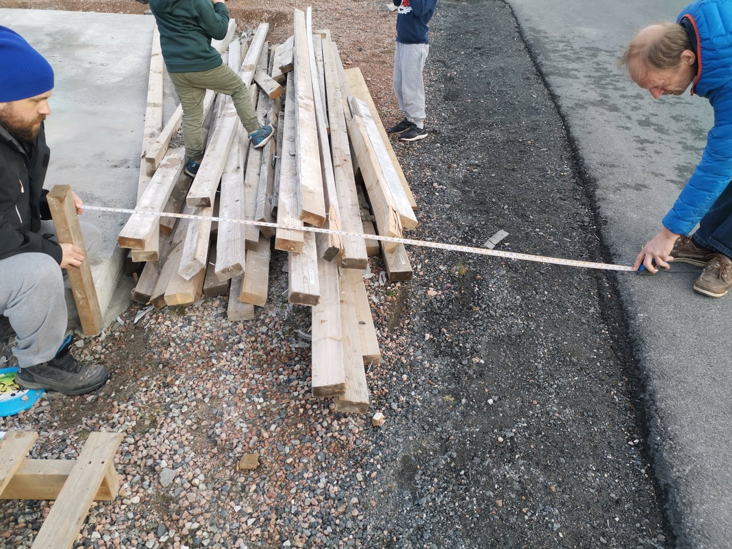 Fotografi av en byggeplass eller et område under forberedelse, der personer måler opp en markering på bakken med en målebånd. Det er også en haug med tømmer ved siden av.