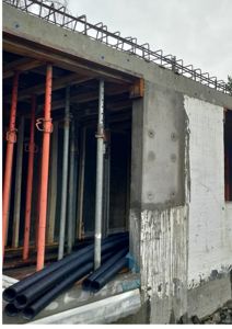 Fotografi av en byggeplass som viser en betongvegg med armering, støttesøyler og rør under et betongtak.