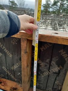 Fotografi av en hånd som holder en målebånd mot en trekonstruksjon under bygging, med isolasjon og takstoler i bakgrunnen.