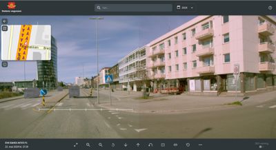 Fotografi av en gatekryssing med bygninger, trafikk og veimarkeringer.