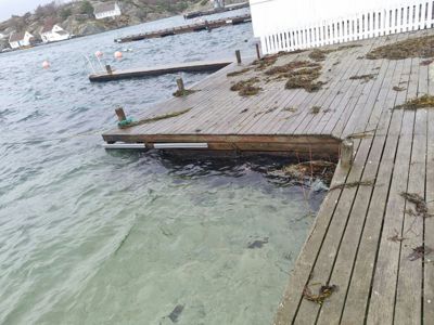 Fotografi av en utendørs scene som viser en treplattform eller kai ved vannet, med en hvit bygning i bakgrunnen.