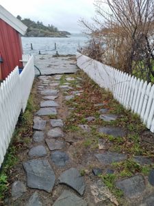 Fotografi av en sti med steinplater som fører fra en rød bygning mot en kai ved vannet, med hvit hekk langs siden.