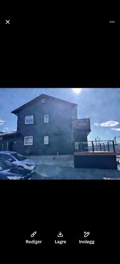 Fotografi av en mørk, to- eller treetasjers bolig sett fra siden, med bil foran og sollys i bakgrunnen.