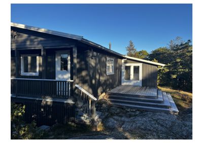 Fotografi av en mørk, trepanelt hytte eller hus sett fra siden. Bildet viser fasaden, vinduer, dører, en veranda med trapper og omgivelsene med trær og stein.