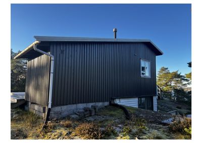 Fotografi av en mørk, paneltett hytte eller uthus sett fra siden. Bygningen står på en skråning med steinmur og har en hvit nedløpsrør.