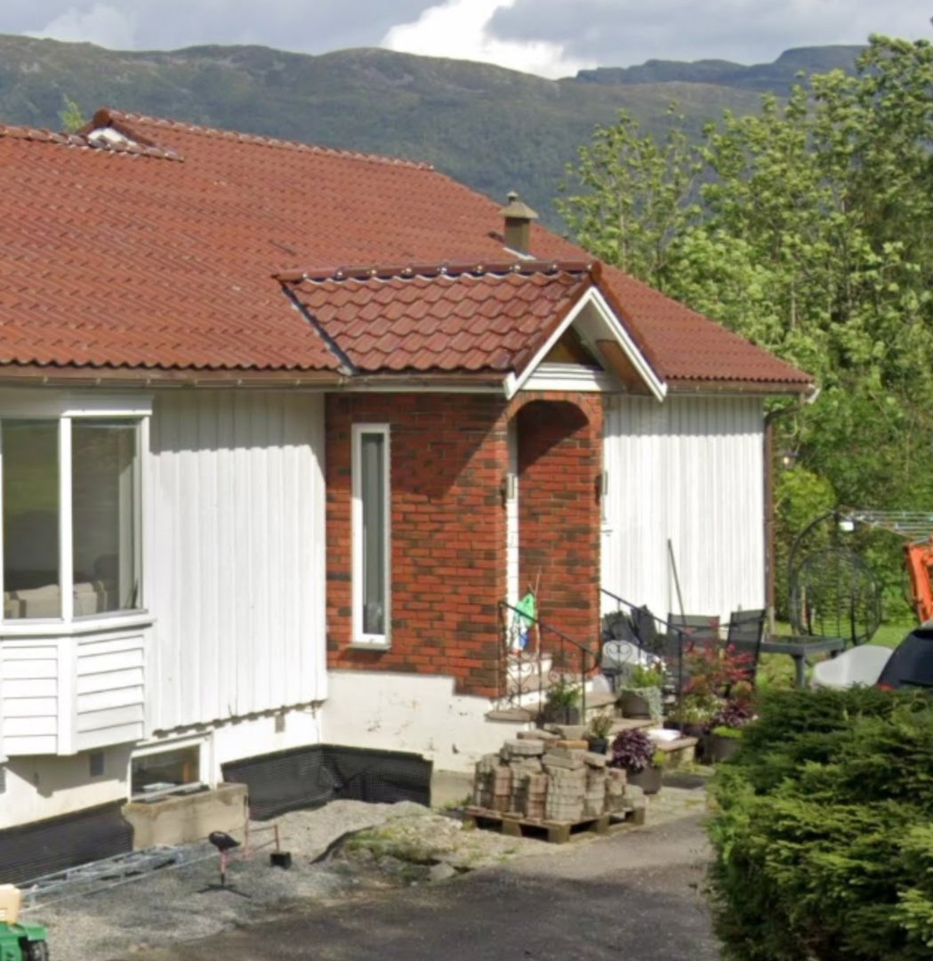 Fotografi av en eksisterende enebolig med hvite panelvegger, rød teglflis tak og en delvis utsatt fasade med murverk. Bildet viser også omgivelser med trær, fjell i bakgrunnen og byggematerialer på pallen.