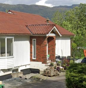 Fotografi av en eksisterende enebolig med hvite panelvegger, rød teglflis tak og en delvis utsatt fasade med murverk. Bildet viser også omgivelser med trær, fjell i bakgrunnen og byggematerialer på pallen.