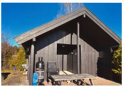 Fotografi av en mørk trehytte eller uthus sett fra fronten, med utemøbler og omgivelser.