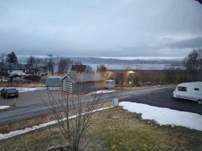 Fotografi av en tomt eller eiendom med en liten uthus, en campingvogn og utsikt over vann og fjell i bakgrunnen.