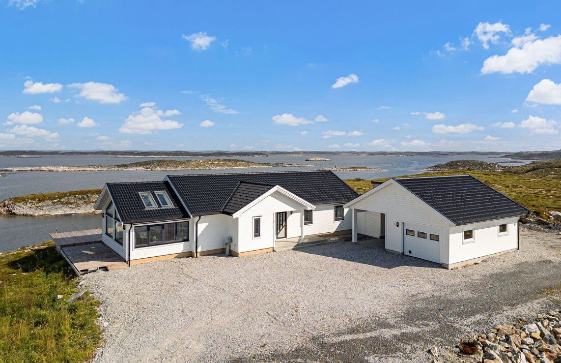 Fotografi av en nybygd hvit villa med svart tak, sett fra en høyde. Bygningen ligger på en grusflate med utsikt over vann og øyer.