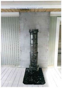 Fotografi av et innvendig utsnitt som viser et antikt ildsted, en gråmalt vegg og gulv med hvite planker.