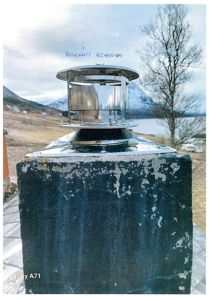 Fotografi av en metallisk røykhatt (merket 'REHODPIPE') montert på en røykpipe. Bakgrunnen viser et landskap med fjell og vann.