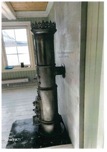 Fotografi av et gammelt, svart jernovn innendørs i et rom med hvite vegger og et vindu.