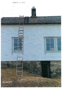 Fotografi av en hvitmalt bygning sett fra siden, med en steinmur i bunnen, et tak med takpanner, en røykpipe og en trapp som leder opp til taket.