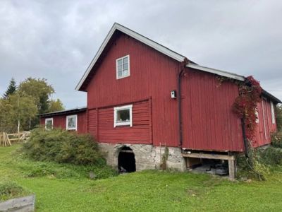 Fotografi av en rød, tradisjonell bygning (muligens et gammelt hus eller uthus) sett fra siden. Bygningen har en steinfundament, vinduer og er omgitt av grønt.