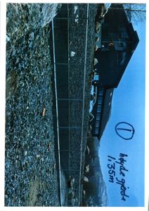 Fotografi av en mørk bygning sett fra siden, med en steinete bakke i forgrunnen og en håndskrevet notis om 'høyde gjerde 1,35m'.