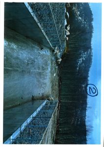 Fotografi av en betongkonstruksjon (kanskje en dam eller vannbeholder) i et naturområde med skog og steiner.