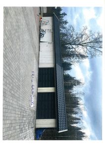 Fotografi av en garasje med hvite vegger og mørke dører, sett fra siden. Bildet viser også en asfaltert eller steinlagt fortau, en sykkel og bakgrunnen med skog og himmel.