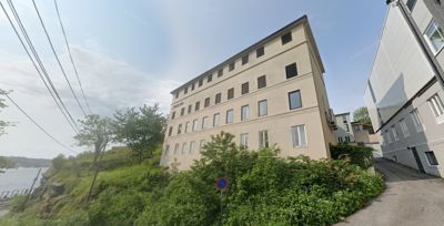 Fotografi av en fleretasjers bygning sett fra en vinkel, med omgivende natur og infrastruktur.