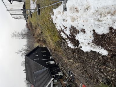 Fotografi av en svart hytte sett fra siden, med snø på bakken og en gjerde i bakgrunnen.