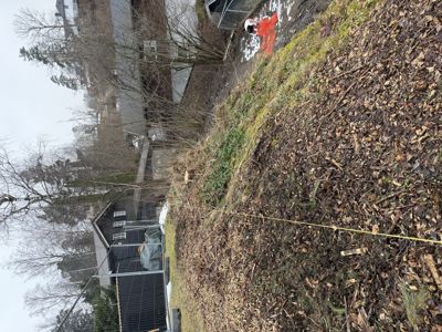 Fotografi av en tomt med et hus i bakgrunnen, der en person i oransje vest står ved en gul målebåndlinje.