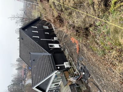 Fotografi av en svart hytte sett fra siden, med omgivende terreng og vegetasjon.