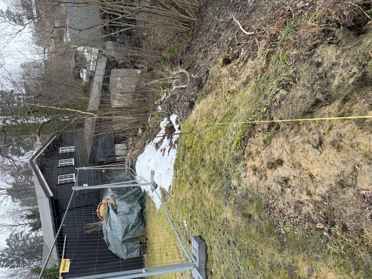 Fotografi av en bygning (svart hus) sett fra siden, med en gjerde i forgrunnen og terrengbakke.