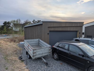 Fotografi av en ferdig eller nesten ferdig garasje med mørke trepaneler og en stor garasjeport. Det er parkert bil og en slippbillett foran.